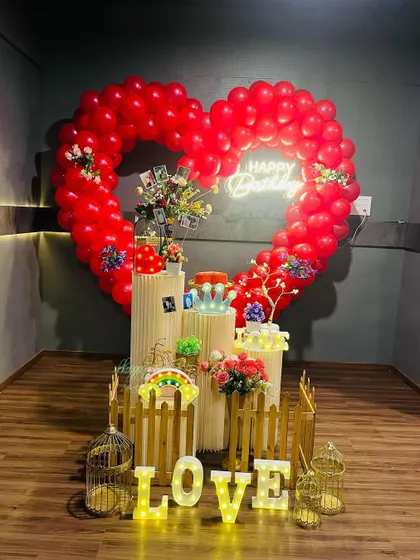 A romantic surprise birthday decoration with a large heart-shaped balloon frame. The setup is filled with personal touches, including photos, light-up "LOVE" letters, and a neon "Happy Birthday" sign.
