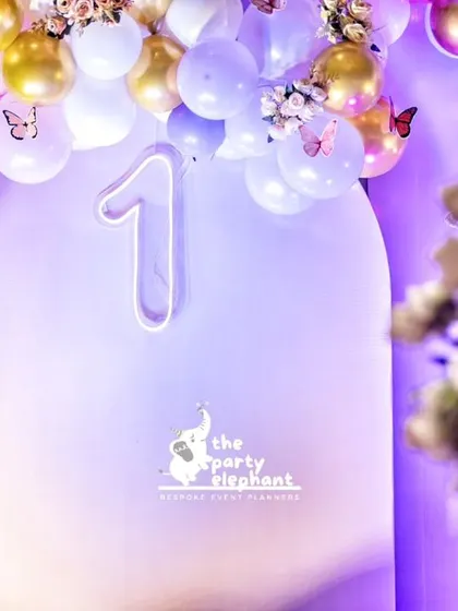 A neon "1" sign glows amidst a cascade of pastel and gold balloons, dotted with delicate butterflies for a modern yet magical touch.