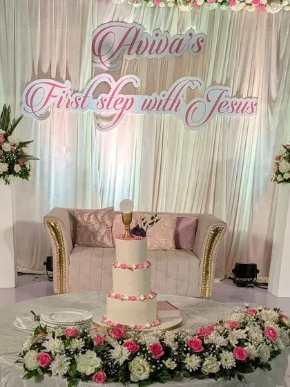 The cake table and stage backdrop for Aviva's 'First step with Jesus.' The design features angelic cutouts, soft draping, and beautiful floral arrangements in a pastel pink and white palette.