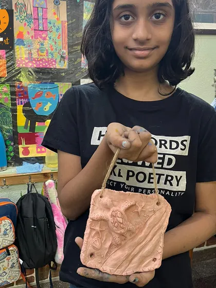 This student proudly displays her finished clay mural. This project involved sculpting shapes and pressing textures into the clay, resulting in a beautiful and personal piece of art.
