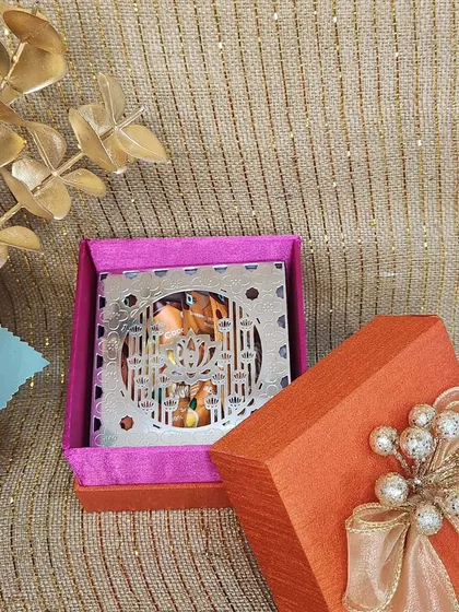 A mini Diwali favor box in bright orange and pink. The intricate silver cutout on top adds a touch of festive sparkle, making it a delightful small gift.