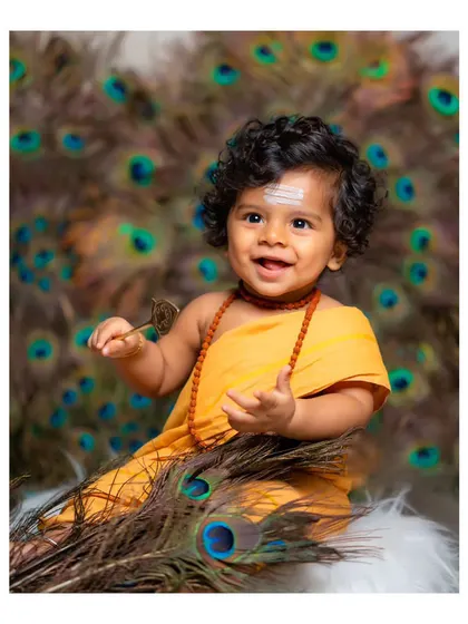 An adorable baby dressed as Lord Murugan (Kartikeya) with a peacock feather background.