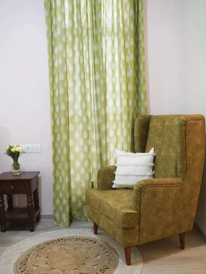 A cozy corner in the guest bedroom, featuring a comfortable wingback chair in a rich green upholstery. The leaf-patterned curtains and jute rug complete the nature-inspired, tranquil vibe of the room.