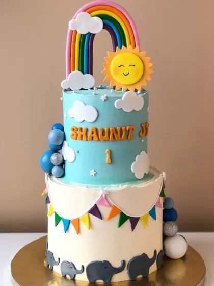A bright and happy two-tiered cake with a sunshine and rainbow theme, and little elephants marching along the bottom.