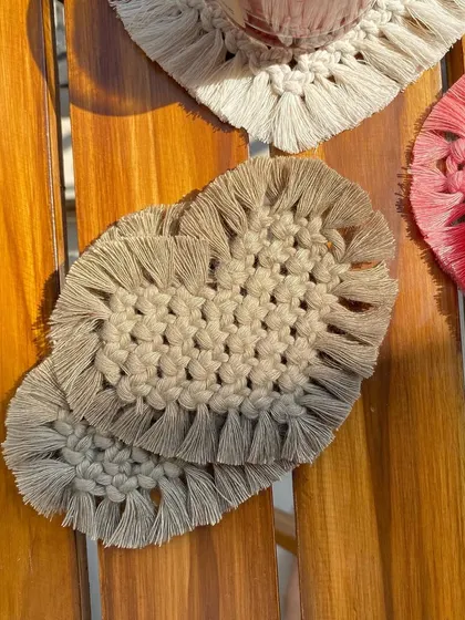 A close-up of the beige macrame heart coasters. The intricate knotting and fringed edge give them a lovely bohemian feel.