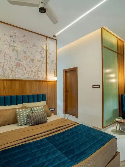 A full view of the master bedroom, highlighting the floral wallpaper panel above the bed, framed by warm wood. The design is balanced and serene, with a cozy reading nook in the corner.