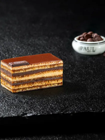 An elegant multi-layered opera pastry, precisely cut and placed on a dark slate, highlighting its glossy top and defined layers for a premium feel.