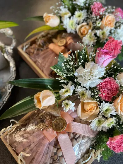 A bespoke Samdhi invitation hamper, featuring a tray of fresh flowers and a box of premium pistachios. The gift is elegantly wrapped with a pink ribbon and a custom wax seal.
