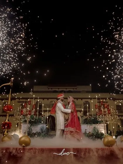 A spectacular jaimala ceremony at a palace, with fireworks lighting up the night sky as the couple exchanges garlands on a stage filled with smoke.
