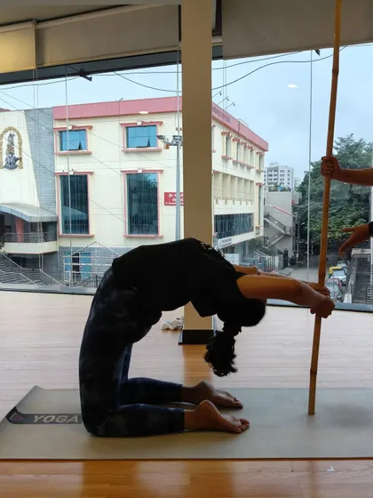 A student uses a stick for support in a deep forward bend from a kneeling position, targeting the spine and shoulders.