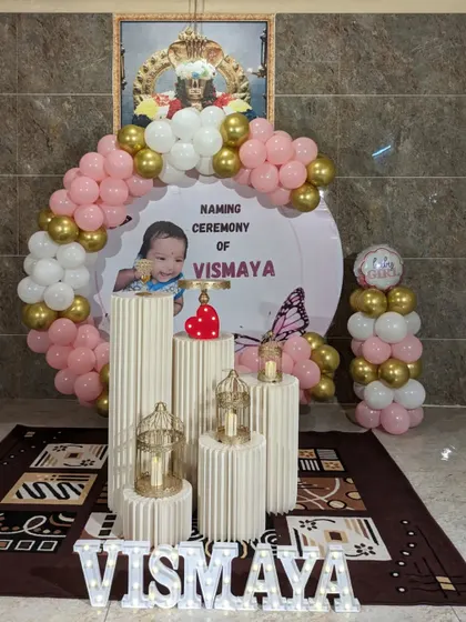 A modern and personalized naming ceremony decor. This setup includes a circular backdrop with the baby's name and photo, a pink and gold balloon arch, and decorative pedestals.