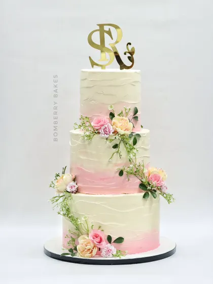 A three-tier buttercream wedding cake with a soft pink ombre effect and adorned with fresh pastel flowers and greenery.