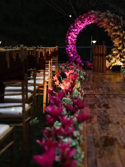 An enchanting evening ceremony setup. The aisle is flanked by vibrant pink lilies leading to a circular arch of flowers and pampas grass, all beautifully illuminated for a romantic nighttime event.