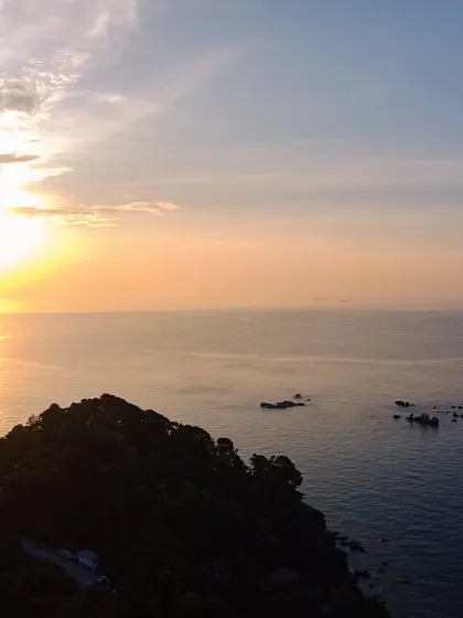 An aerial view of the sun setting over the ocean off the coast of Bintan Island, with the silhouette of a hill in the foreground.