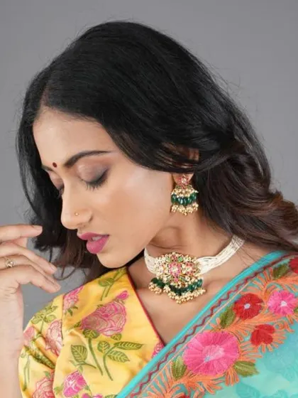 A close-up of the green and ruby choker set on a white beaded base. The matching jhumka-style earrings feature the same floral motif and color palette.