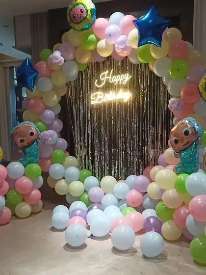 A colorful Cocomelon-themed birthday decoration. I create a circular balloon arch in pastel colors, with a silver tinsel curtain, a 'Happy Birthday' neon sign, and Cocomelon character balloons.