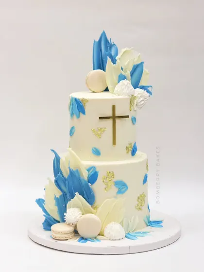A two-tier baptism cake in white and blue, decorated with abstract chocolate sails, macarons, and a simple gold cross.