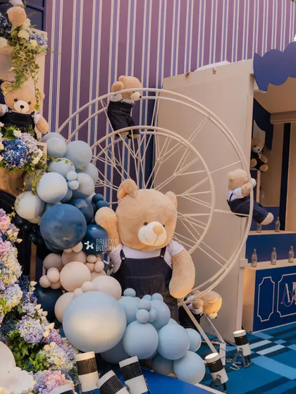 Teddy bears climb a Ferris wheel next to a cascade of blue and white balloons. This playful detail adds a dynamic element to the overall birthday party decor.