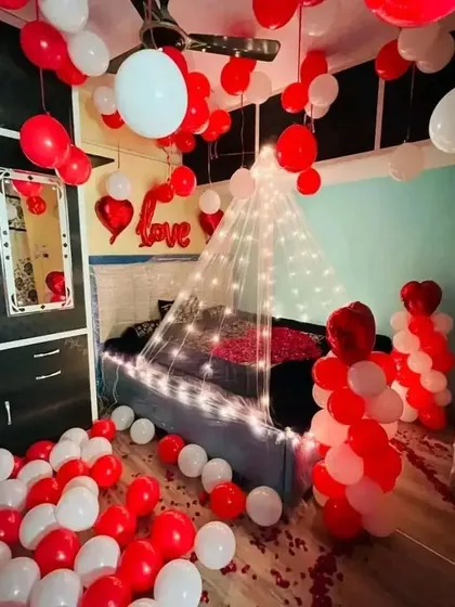 A simple yet romantic bedroom decoration. The setup includes red and white balloons on the floor and bed, a "LOVE" foil balloon, and a light-up canopy.