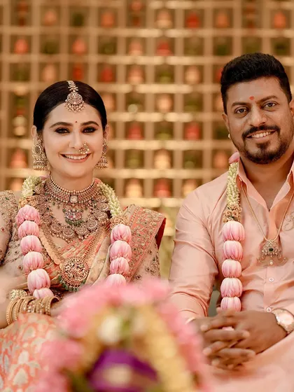 A happy couple posing during their naming ceremony. Their coordinated peach-toned outfits, a silk saree for her and a silk shirt for him, create a warm and joyful picture.
