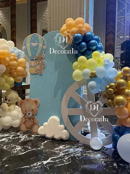 A cozy photo corner with a large wheel prop, teddy bears, and hot air balloon cutouts, all tied together with a blue, yellow, and orange balloon garland.