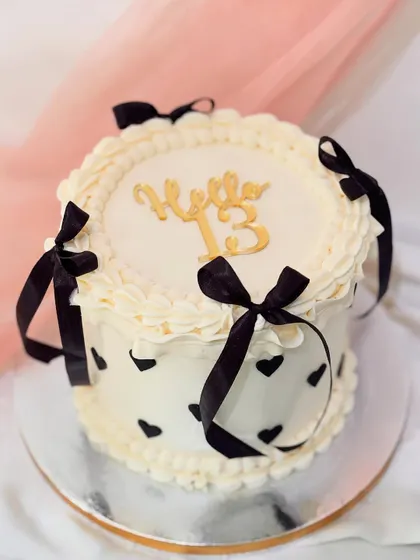 A chic vintage-style cake for a 13th birthday. The design features white buttercream ruffles, black heart details, and is finished with elegant black ribbons.