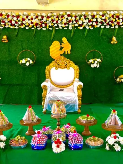 A full view of a baby shower stage, featuring a lush green backdrop, a central throne, and a beautiful arrangement of traditional sweets and offerings in the foreground.