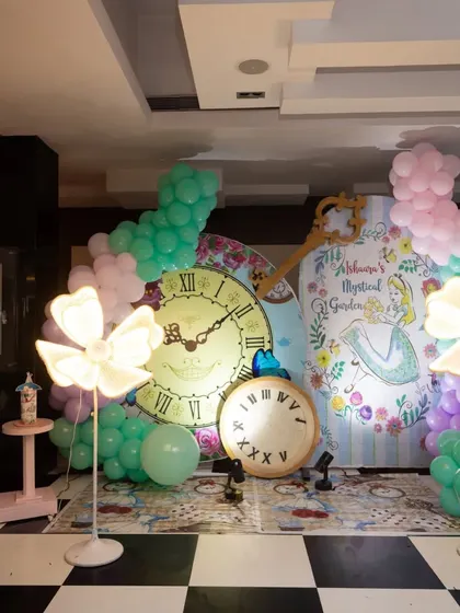 A mystical 'Alice in Wonderland' photo booth. The backdrop features giant clocks and a hand-drawn Alice, framed by glowing flower lights and whimsical balloon garlands.