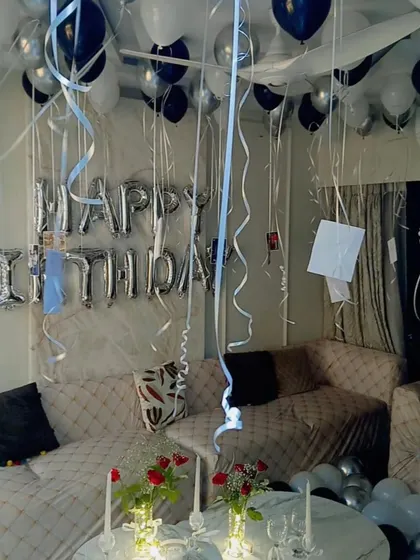 A full view of a surprise birthday room decoration, with silver and black balloons on the ceiling, hanging photos, and a beautifully arranged table for two.