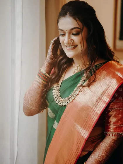 A lovely candid portrait of the bride. She adjusts her jewelry by the window, bathed in soft, natural light that highlights her smile and traditional green and orange saree.