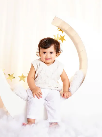 A smiling baby sitting on a white crescent moon prop, dressed in an adorable all-white outfit. A heavenly and sweet portrait.