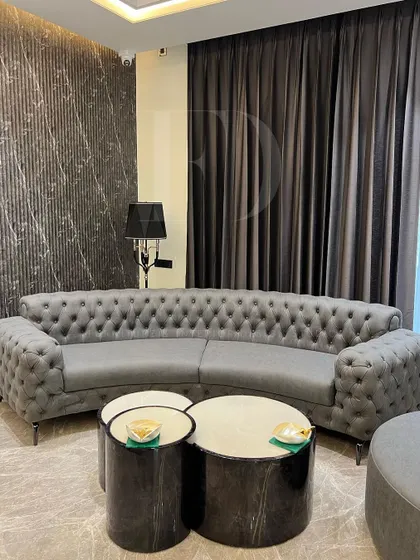 This living room features a curved, tufted Chesterfield sofa in grey, paired with a matching circular ottoman. The set creates a cohesive and elegant look, grounded by dark marble nesting tables.