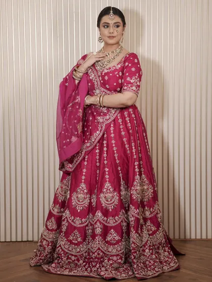 A full view of a bride in a deep magenta lehenga. The outfit is paired with a matching Kundan necklace set, showing a complete and cohesive look available for rent.