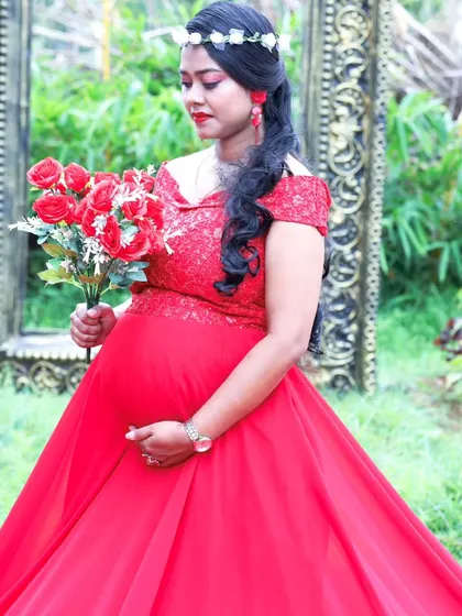An elegant maternity photo of a woman in a red trail gown, holding a bouquet of red roses in a garden.