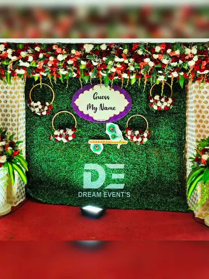 A full stage view for a naming ceremony, featuring a lush green backdrop, a floral valance of red and white roses, and a playful "Guess My Name" sign with Krishna flute details.