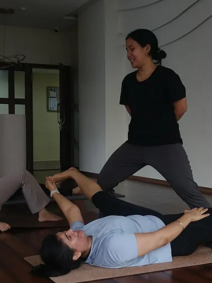 An instructor guides a student through a supine hip opening stretch. These immersive practices focus on releasing deep seated tension in the hips for greater mobility.