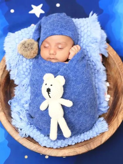 A classic sleeping pose in a wooden bowl. The baby is holding a small teddy bear, perfectly swaddled in blue, creating a timeless and adorable newborn photograph.