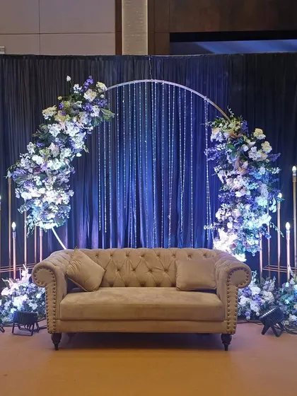 A modern and chic reception stage featuring a circular arch with blue and white florals and elegant crystal hangings.