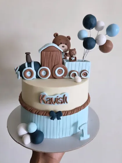 All aboard the birthday express! This charming two-tiered cake features a teddy bear driving a train, complete with a balloon bouquet. It's a delightful theme for a first birthday celebration.
