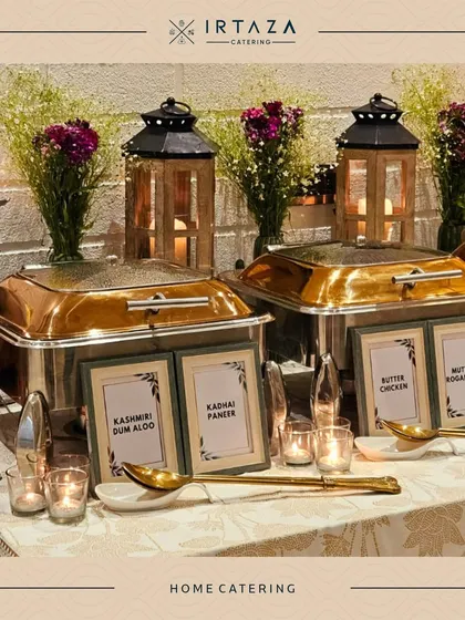 A classic home catering arrangement with silver chafing dishes labeled with "Kashmiri Dum Aloo," "Kadhai Paneer," and more. The wooden lanterns and baby's breath flowers add a rustic yet elegant touch.