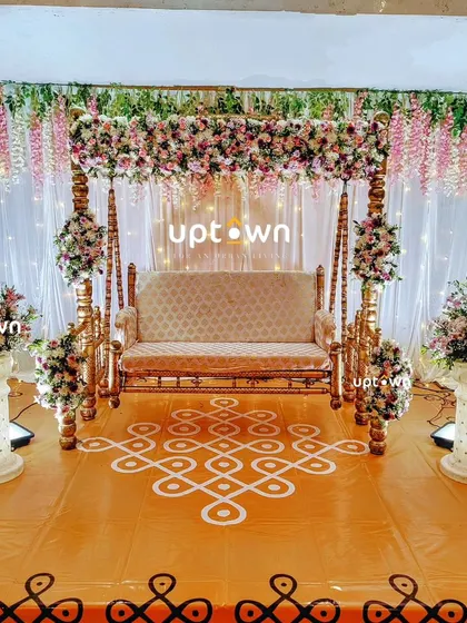 I specialize in traditional baby shower decorations that feel both classic and beautiful. This setup features an ornate wooden swing decorated with hanging flowers, set against a backdrop of white drapes and a traditional kolam floor design.