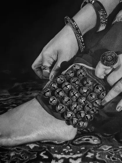 A complete view of the dancer's foot as she secures the ghungroo, set against a patterned carpet. The entire scene is brought to life through careful shading.