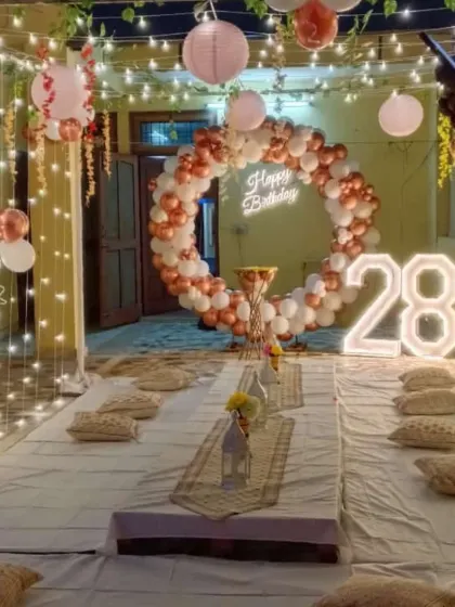 An intimate terrace party setup for a 28th birthday. The decoration includes a rose gold and white balloon ring, a neon sign, and a canopy of fairy lights over a low seating arrangement.