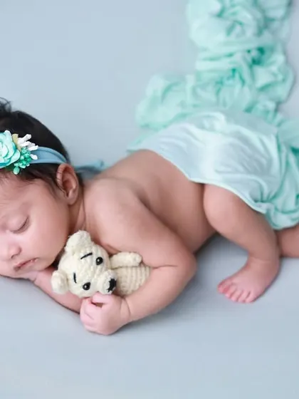 A sleeping baby in a side-lying pose, hugging a tiny teddy bear. The mint green wrap and headband complete the look.