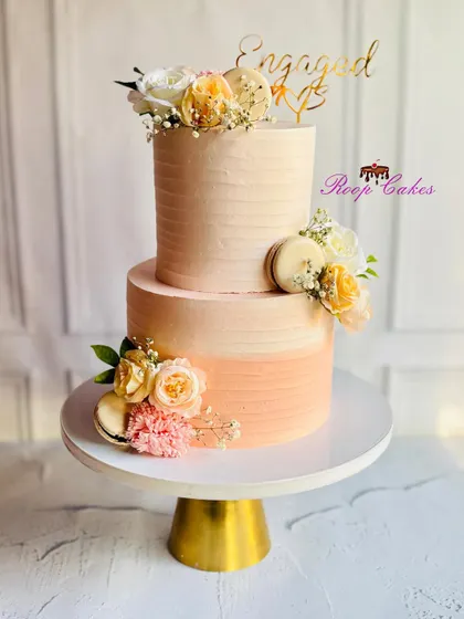 A sweet beginning to forever. This two-tier engagement cake features a warm peach ombre effect, adorned with fresh flowers and macarons for a touch of modern elegance.