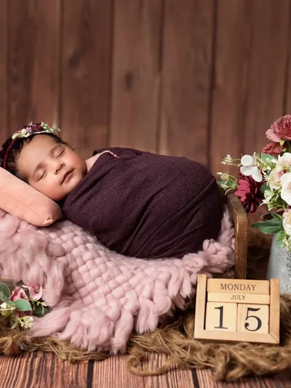 A sleeping newborn girl wrapped in a deep purple swaddle, resting peacefully in a tiny bed with floral accents.