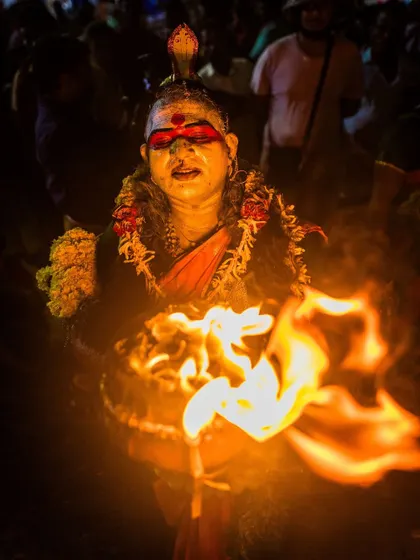 A performer holds a flaming vessel, their face painted and expressive. The firelight creates a dramatic, glowing effect that highlights their features.