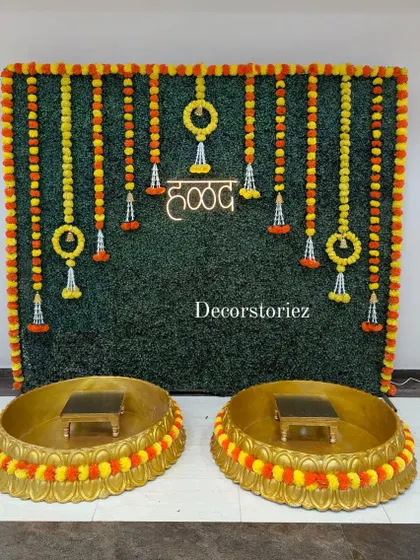 We designed this complete Haldi ceremony setup featuring a lush artificial grass wall backdrop, traditional marigold garlands, and a bright neon 'हळद' sign. The two large golden Urlis and matching patlas (stools) are also available from us on a rental basis.