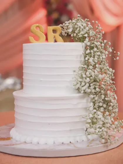 A classic two-tiered white wedding cake with a textured buttercream finish. It's personalized with the couple's initials "SR" and a cascading arrangement of fresh baby's breath for a timeless look.