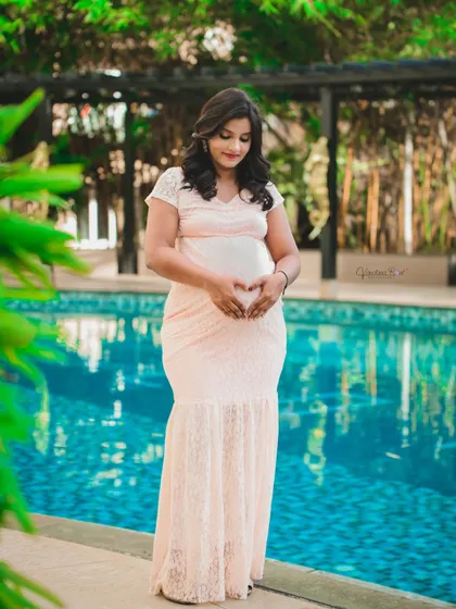 A serene solo portrait of a mother-to-be in a delicate lace gown, making a heart shape with her hands over her belly by a tranquil pool.